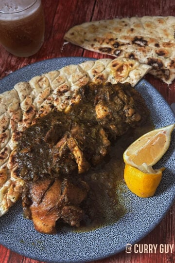 Black chicken curry on a serving plate with a naan and lemon wedges.